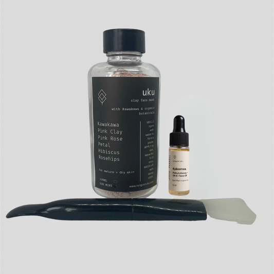 Set of skincare products including a bottle of Kawakawa and Pink Clay face mask powder, 5ml dropper bottle of Kokomea face oil, and spatula on a light gray background