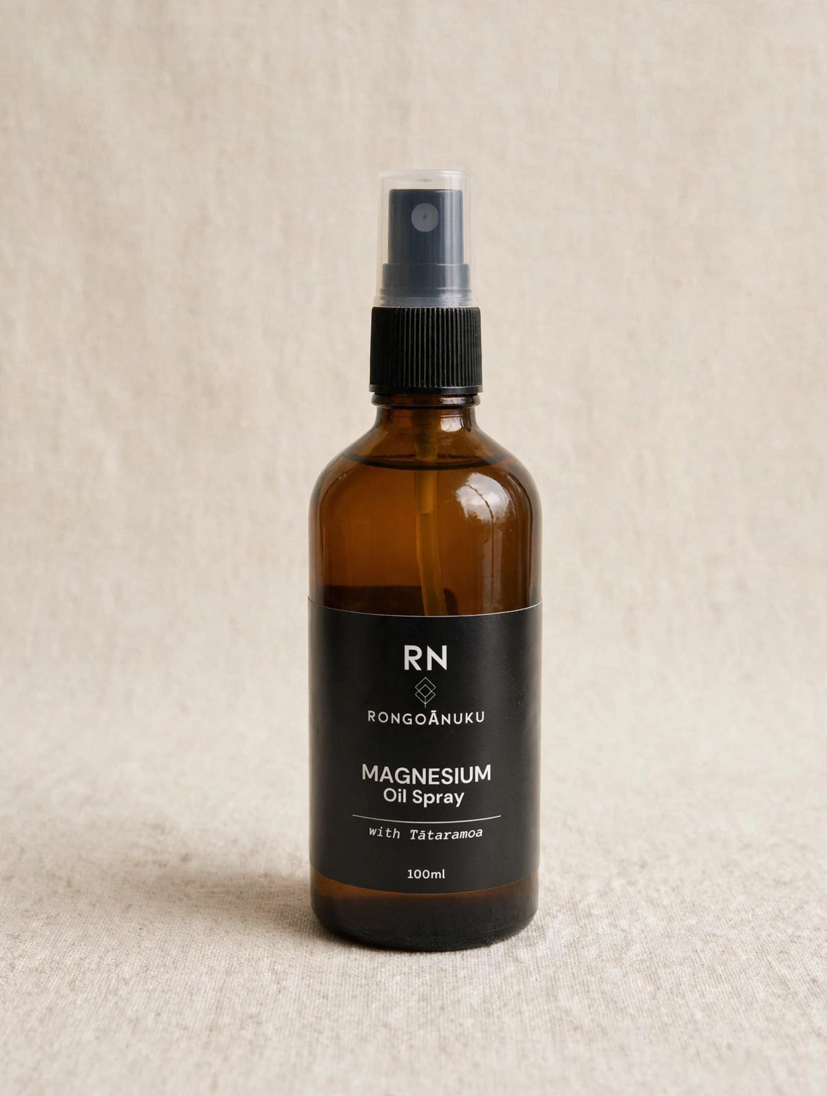 An amber glass spray bottle of Rongoanuku Magnesium Oil Spray with a black label and spray nozzle, set against a neutral beige textured background.